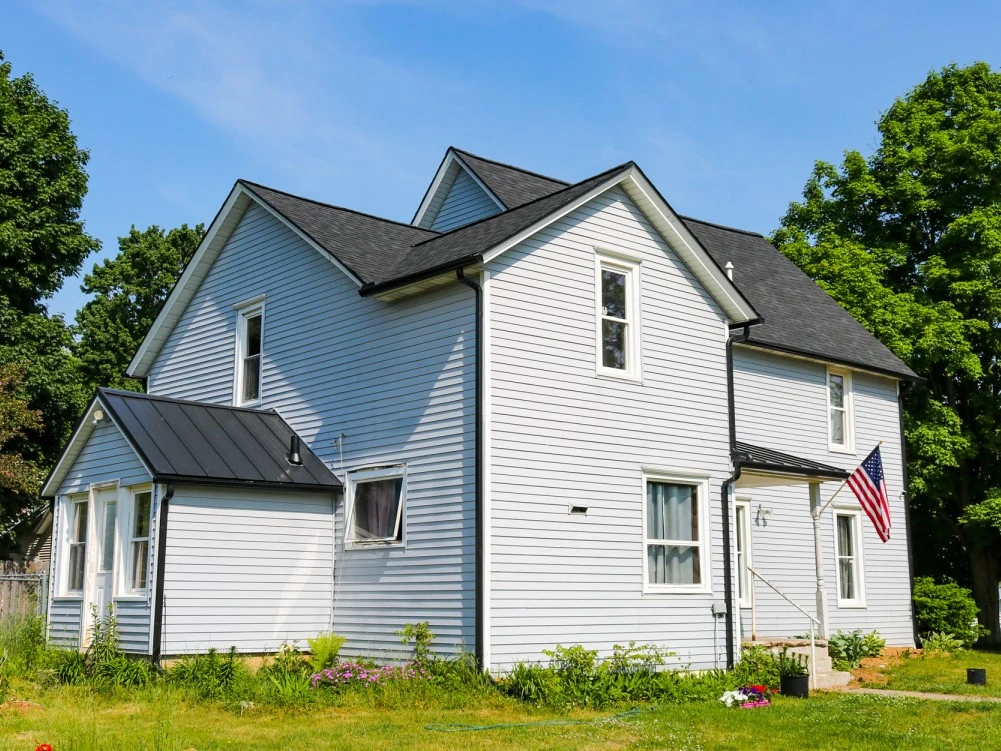 A two-story American suburban home with light blue vinyl siding and a black metal roof on a sunny day. The house features multiple windows, a small front porch, and an American flag displayed on a flagpole. The yard is partially landscaped with a lawn, a few flowerbeds, and surrounded by mature trees.