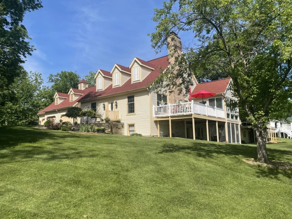Spacious multi-story house with dormer windows and expansive decks surrounded by lush greenery and mature trees on a sunny day.