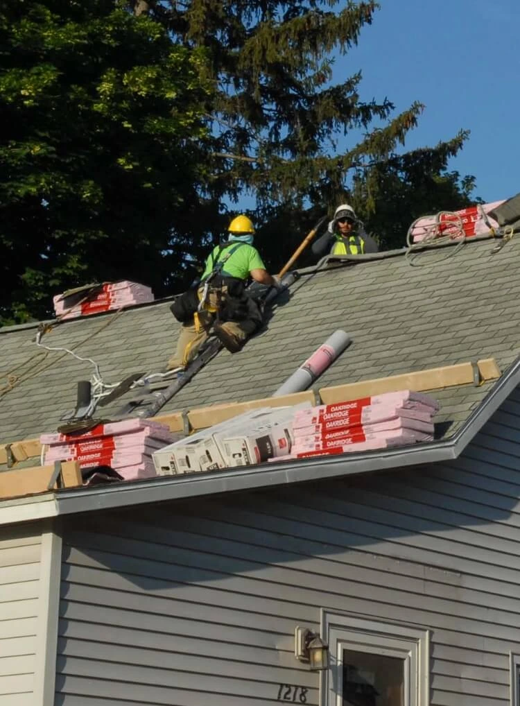 Roofing professionals wearing safety gear work on a sloped residential roof during a roof replacement, with new shingles and materials staged along the roofline.